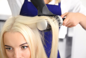 Fototapeta premium Professional hairdresser blow drying blonde woman's hair with round brush in a modern salon setting, the stylist uses a hairdryer and brush to create a smooth, styled look