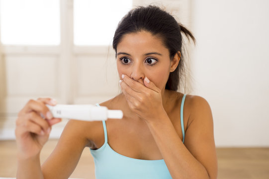  latin woman holding pregnancy test on bed at home looking at positive result in shock and stress
