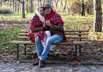 Young couple sitting on bench at the park covered in plaid/blanket. Autumn season.
