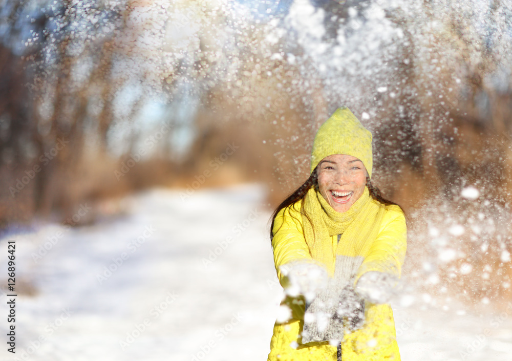 Wall mural Winter snow fight happy girl throwing snow playing outside. Joyous young Asian woman having fun in nature forest park on snowy day wearing yellow outerwear with warm accessories: gloves, hat, scarf.