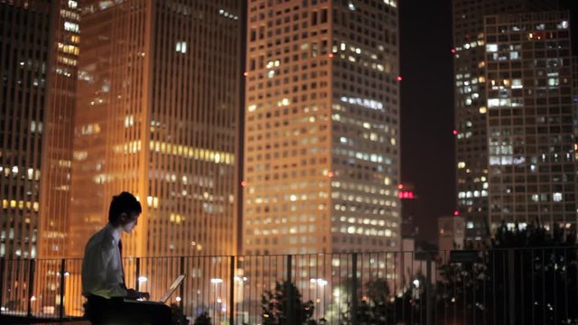 WS Businessman sitting on terrace using laptop, cityscape in background at night / Beijing, China