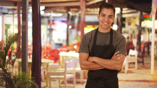 Happy handsome young Latino waiter standing in outdoor patio of restaurant smiling. Friendly millennial server wearing apron while waiting tables at small business