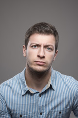 Moody portrait of distrustful young executive man in blue shirt looking at camera over gray background