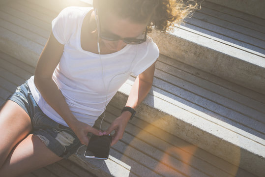 View from above. Sunny summer day, the girl in sunglasses, white T-shirt and denim shorts sitting on the stone steps and listening to music, while holding a smartphone.