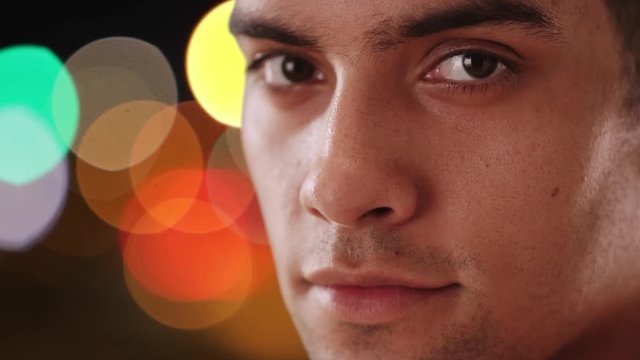 Close up portrait of Latino man looking at camera on city street at night. Hispanic millennial in 20s with bokeh lights behind him