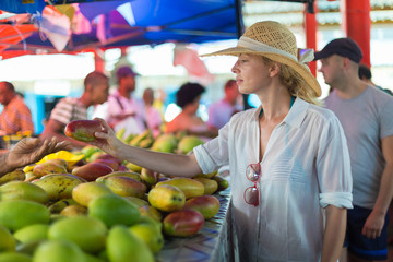 Female traveler wearing elegant colonial style white tunic and hat buying fresh tropical fruit on...