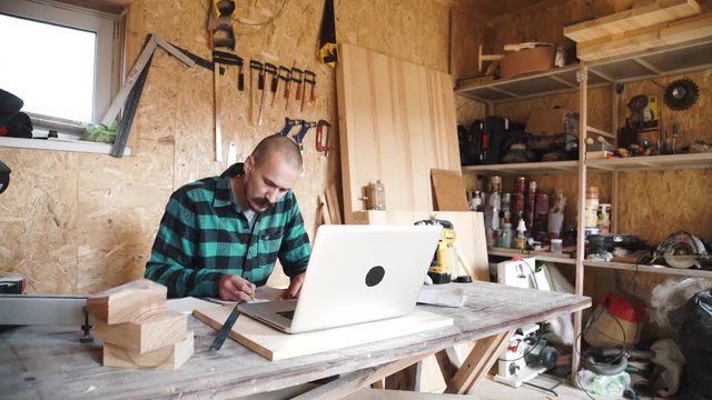 Portrait of bold moustache retired carpenter sitting at his workshop and working with laptop. Small business.