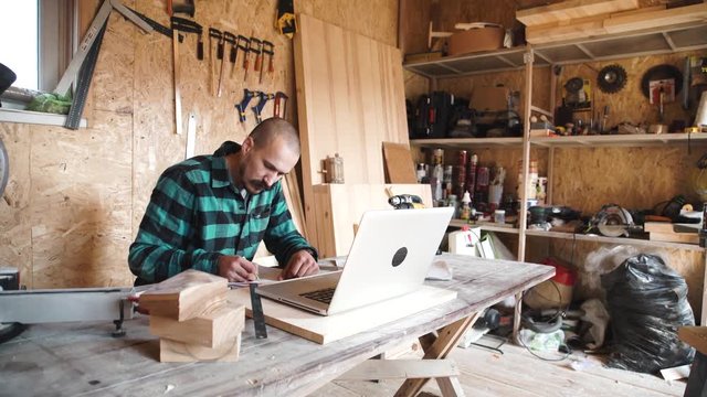 Portrait of bold moustache retired carpenter sitting at his workshop and working with laptop. Small business.