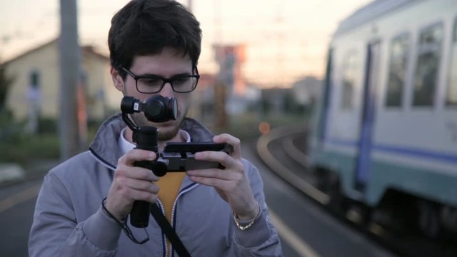 Young boy Glasses with a Grey Jacket is Taking a Video Footage with his modern Gimbal Camera Linked with his Smartphone.