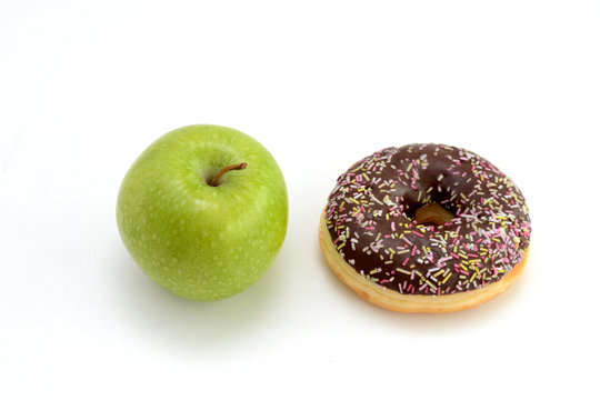 Close-up of tasty chocolate donut and fresh green apple on white background suggesting healthy eating concept