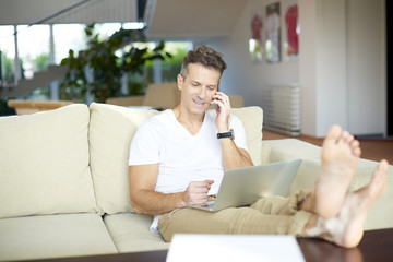 Working at home. Full length shot of a casual middle aged man making call and working on laptop while sitting at home. 