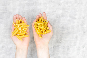 Obraz premium Handful of uncooked pasta flat lay. Female hands holding heap of raw fussilli and penne noodles on white canvas background