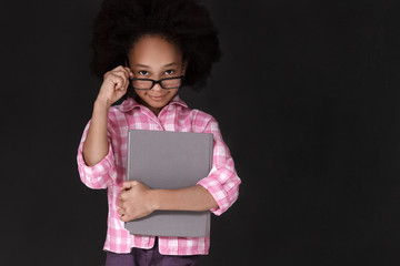 Cheerful girl holding book and adjusting glasses while standing against blackboard