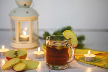 Чай в стеклянной кружке с долькой яблока и кусочками яблока на белом столе. Стол украшен свечами, яблоками, еловой веткой, желтой салфеткой и фонарем со свечой