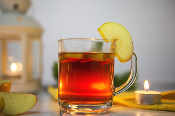 Чай в стеклянной кружке с долькой яблока и кусочками яблока на белом столе. Стол украшен свечами, яблоками, еловой веткой, желтой салфеткой и фонарем со свечой