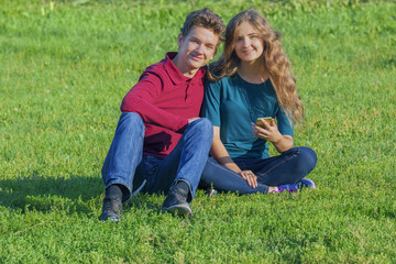 Fototapeta premium Couple teenagers sitting on the green lawn with a smartphone