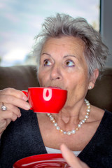 Portrait of a senior lady enjoying a cup of coffee from a bright red cup and saucer that she is holding in her hands