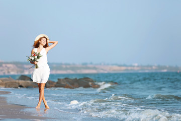 Fototapeta na wymiar Young happy woman with bouquet of flowers walking along seashore