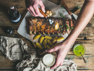 Man eating roasted pork ribs with garlic, rosemary and green herb sauce on rustic wooden table. Man' s hands holding fork with fried potato and glass of dark beer, top view