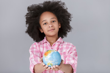 World in my hands! Cheerful little girl holding a globe while standing against grey background