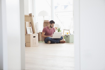 Young man in new flat with cardboard boxes using laptop