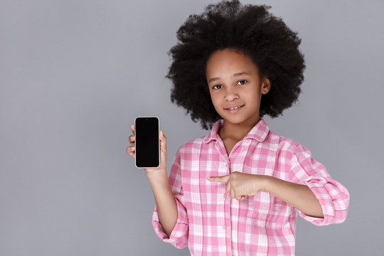 Copy space on her phone. Pretty little girl holding mobile phone and pointing at it while standing against grey background