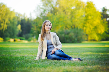 Pensive beautiful young woman dressed in a lightweight business style, resting, sitting on the grass, gaze to the side, relaxed posture, full growth.