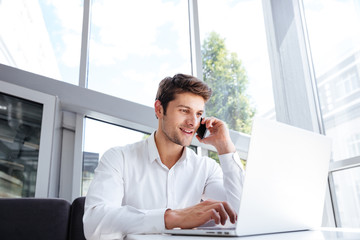 Happy young businessman talking on cell phone and using laptop