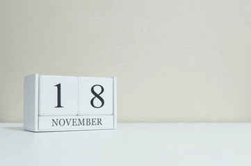Closeup white wooden calendar with black 18 november word on blurred white wood desk and cream color wallpaper in room textured background with copy space , selective focus at the calendar