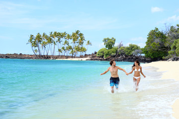 Fototapeta na wymiar Happy couple laughing together holding hands running having fun splashing water in the ocean waves. Young beautiful fit slim people enjoying their happy lifestyle in paradise destination beach.