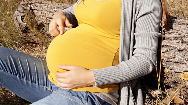 Unknown woman sits leaning on a log and caress her pregnant belly. Focus to the belly and hands