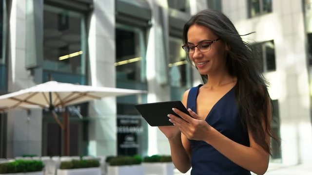 Beautiful young lady in glasses holding a tablet on hands, walking on the street.