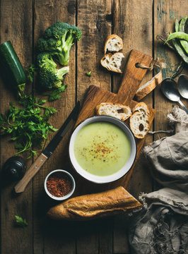 Homemade pea, broccoli, zucchini cream soup in white bowl with fresh baguette on wooden board over rustic background, top view, vertical composition