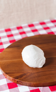 Single water buffalo mozzarella mozzarella di buffala cheese ball on wooden cheese chopping board on red and white checkered tablecloth with brown burlap background with copy space above