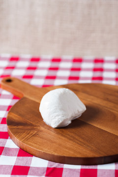 Single water buffalo mozzarella mozzarella di buffala cheese ball on wooden cheese chopping board on red and white checkered tablecloth with brown burlap background with copy space above