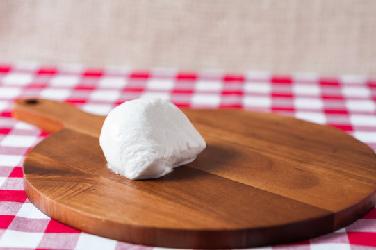 Single water buffalo mozzarella mozzarella di buffala cheese ball on wooden cheese chopping board on red and white checkered tablecloth with brown burlap background with copy space above