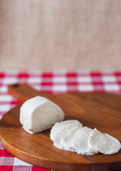 Fototapeta na wymiar Water buffalo mozzarella mozzarella di buffala cheese in ball and sliced on wooden cheese chopping board on red and white checkered tablecloth with brown burlap background with copy space above