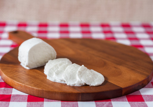 Water buffalo mozzarella mozzarella di buffala cheese in ball and sliced on wooden cheese chopping board on red and white checkered tablecloth with brown burlap background with copy space above
