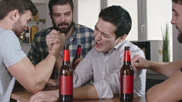 Asian man winning male friend in arm wrestling while drinking beer with one hand