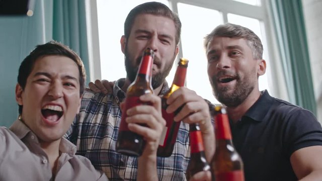 Male friends drinking beer and clinking bottles in celebration while taking selfie at party