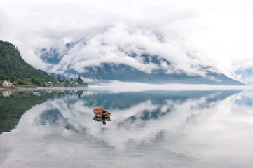Morning by the fjords, clouds on the mountains with reflection on the water and small boat, Norway