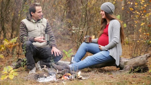 Beautiful  pregnant woman with her husband resting in the forest.  They sit near the fire, drink hot tea talking and smiling. The concept of family happiness
