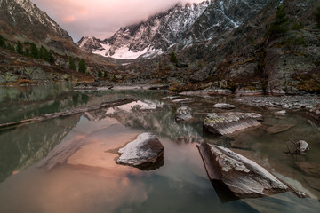 Mountain pass Kuyguk hide by the clouds colored by the sunset. In the foreground are drift wood, stones and rocks covered with lichen. They are in smooth and clear water of Lake Kuyguk, Altai
