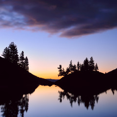 Twilight. Mountain slopes and larch trees silhouette is reflected in the smooth surface of the lake Kuyguk. Clouds backlit by the sunset, Altai