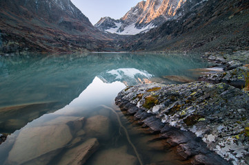 Huge rock with large crevices covered by lichen is in a smooth transparent water of turquoise mountain lake Kuyguk. Calm water reflects slopes and large glacier on the way to the mountain pass, Altai