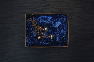 Box with Christmas decorations on a black wood background. Vintage New Year's toys. Blue wrapping paper and kraft box. Top view, flat lay