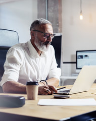 Adult professional businessman wearing a classic glasses and working at the wood table in modern coworking studio.Stylish bearded middle age man using laptop on workplace. Vertical,blurred.