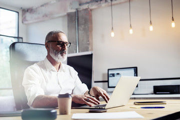 Middle age financial analyst wearing a classic glasses and working at the wood table in modern interior design office.Stylish bearded businessman using laptop on workplace. Horizontal,blurred.