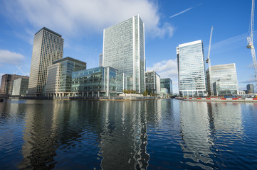 Fototapeta na wymiar London skyline of the modern business complex of Canary Wharf reflecting on calm waters next to construction cranes under bright blue sky