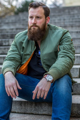 M&auml;nnlicher b&auml;rtiger Hipster in Jeans sitzt auf Treppe und schaut seitlich (nah)